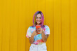 © VISTA by Westend61 - Smiling young woman wearing tie dye t-shirt using smart phone in front of yellow wall