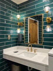 A bathroom with green subway tile walls, gold lights, marble sink and a gold faucet.