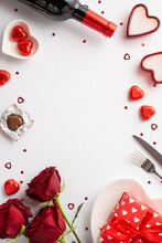 Valentine's Day Concept. Top View Vertical Photo Of Wine Bottle Bouquet Of Roses Giftbox Heart Shaped Saucer Candies Cutlery Candles Confetti On Isolated White Background With Copyspace In The Middle