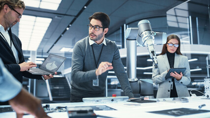 Fototapete - Industrial Robotics Engineer Talking and Discussing the Development of a Robotic Arm Machine with His Colleagues. Scientists with Laptop and Tablet Computers Research the Possibilities of the Robot.
