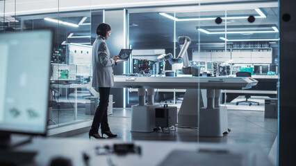 Poster - Industrial Robotics Engineer Standing Next to a Robotic Arm in a High Tech Factory. Female Scientist Uses Laptop Computer to Manipulate and Program the Robot to Pick Up and Move a Microchip.