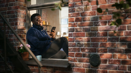 Poster - African American Young Man in Comfortable Casual Clothes Using Smartphone and Sitting on his Windowsill. Black Male Staying Connected and Using Internet in a Brooklyn Style Brownstone House