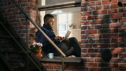 Poster - Black Young Male Smiling and Using Laptop While Sitting on a Windowsill. African American Businessman Enjoying the Weather While Working from Home. He Is Thinking and Answering his Business Emails.