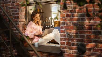 Poster - Young Caucasian Woman Working from Home and Using Laptop While Sitting on Bedroom Windowsill on a Warm Sunny Day. Female CEO Remotely Working on Project in a Spring Atmosphere.