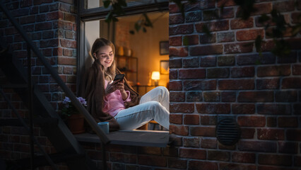 Poster - Beautiful White Woman Using Smartphone While Sitting on an Open Windowsill. Attractive Girl with Healthy Creative Lifestyle Relaxes at Home in the Evening, Spending Some Alone Self-care Time