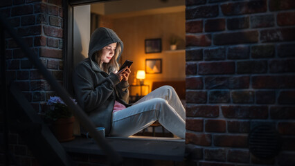 Leinwandbilder - White Woman Wearing a Hoodie Using her Smartphone While Sitting on Windowsill at Night. Female Gamer Playing Addictive Mobile Phone Games Instead of Sleeping. Bad Case of Unhealthy Gaming Addiction