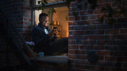 Poster - Black Man Using Smartphone for Online Shopping While Sitting on his Windowsill at Night. e-Commerce Concept of Easy Purchasing, Buying, and Ordering on Website and the Internet.