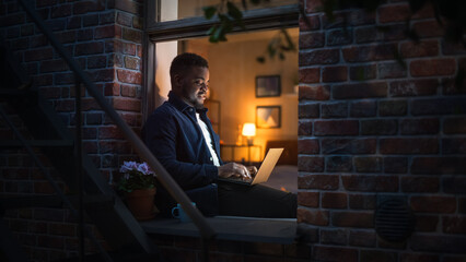 Poster - Young Handsome Black Man Working from Home on Laptop Computer in Windowsill of a Cozy Stylish Loft Apartment. Creative Male Smiling, Checking Social Media, Browsing Internet During Night