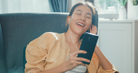 Wall Mural - Close-up Youth asia female girl or university student wear earphone hold smartphone sit on sofa chair take a rest chill listen music singing after work in living room at house. Home lifestyle concept.