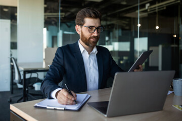 Male office manager in suit sitting at workdesk, taking notes while attending business webinar online