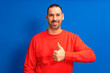 © Andres - Bearded latin man wearing a red sweater raising his thumb up in approval while smiling at the camera in a friendly attitude isolated over blue background.