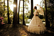 © Cavan Images - A young woman poses in a Southern Belle outfit , including a large, ornate period-dress and parasol, near a lake.