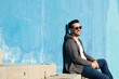 © Cavan Images - Portrait of stylish man sitting on stairs leaning on a blue wall while looking away and smiling