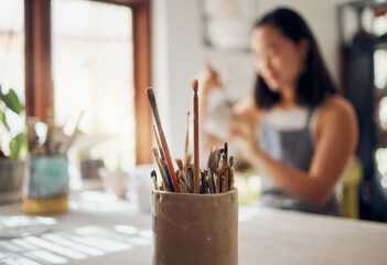 Wall Mural - Paintbrush, art and equipment with a jar on a table in a pottery workshop or studio and a designer woman in the background. Zoom, paint and creative with a female potter working on a design