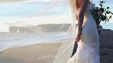 Long Wedding Bridal Veil Flowing In The Wind Behind Caucasian Bride In White Dress Standing On Black Sand Beach During Sunset. Slow Motion Clip.