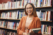 Face, student and woman in university in library ready for learning. Portrait, education and happy female from Brazil standing by bookshelf with book for studying, knowledge and literature research.