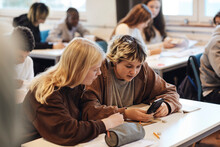 Teenage Girl Sharing Smart Phone With Blond Female Friend Sitting At Desk In Classroom