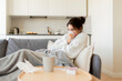 © deniskomarov - A ill girl coughs and uses nasal wipes while sitting on the sofa and wrapped in a duvet. Young woman with symptoms of a viral flu infection is sad at home