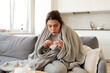 © deniskomarov - Ill young woman takes pills sitting at home on the sofa under a warm blanket