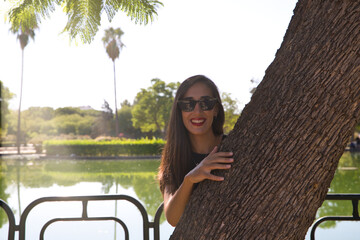 beautiful brunette spanish woman leaning against a tree. In the background you can see a lake in the park. The woman is dressed in elegant black clothes and wears sunglasses. f