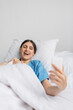© LIGHTFIELD STUDIOS - Positive woman in patient gown using cellphone on bed in hospital ward.