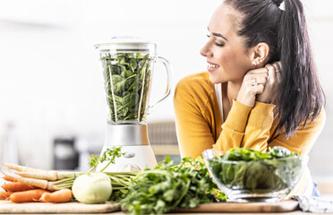 A young woman looks at a blender full of spinach, fresh vegetables around her