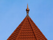 pointy spire on turret with hexagonal sloped building roof top. red clay tile roof. copper spike or spire at the tip. ridge tiles. blue sky background. building materials and construction concept.
