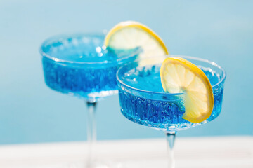 A glass of delicious blue cocktail Blue Lagoon and yellow lemons on the background of the pool. Alcoholic cocktail juicy fruit blue with curacao liqueur, ice cubes and a slice of lemon.