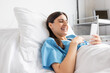 © LIGHTFIELD STUDIOS - Cheerful patient in gown using smartphone while lying on bed in clinic.