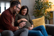 © Dorde - Happy couple feeling movements of a baby in a belly of the expectant mother, excited about a new family member while sitting on a couch and relaxing at home.
