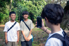 A Boy With A Mobile Phone Taking A Picture Of Two 13 Year Old Boys Side By Side, Outdoors In A Park In Summer.