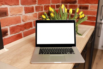 Business, freelance, blogger concept. Laptop with empty white screen on wooden table against brick wall with yellow tulips in vase at home office. Mock up, copy space. Social media, website template