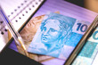 © Rmcarvalhobsb - Notas do Real Brasileiro na tela de um celular. Um caderno e uma caneta na composição. Conceito de economia brasileira e o Real Digital.