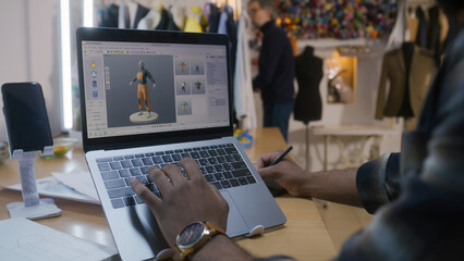 African American fashion designer creates clothes in 3D modeling software on laptop using digital tablet and stylus. He works in atelier workshop. Concept of fashion and technologies in business.