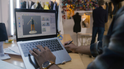 African American fashion designer creates clothes in 3D modeling software on laptop using digital tablet and stylus. He works in atelier workshop. Concept of fashion and technologies in business.