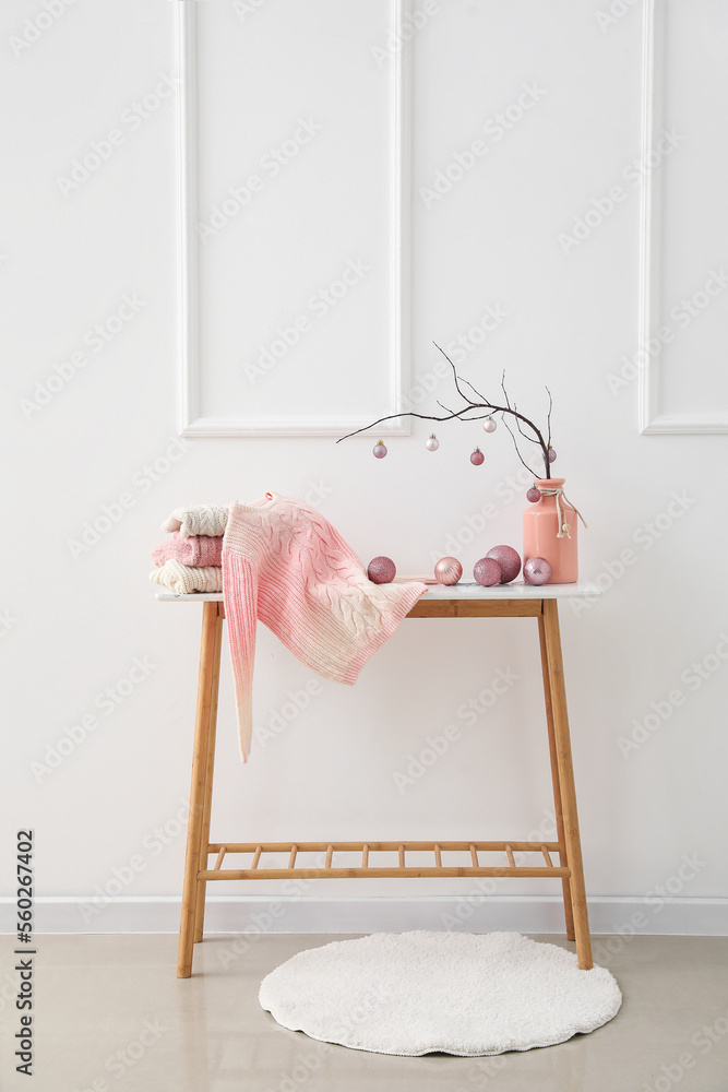 Vase with tree branches, Christmas balls and sweaters on table near light wall