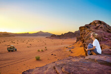 A Woman Sits On A Rocky Mountain Top Watching The Sun Set In The Wadi Rum Desert, A Truck Is In The Distance