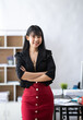 © crizzystudio - Portrait of successful confident pretty, young Asian businesswoman or freelancer standing near work desk in office, wearing formal stylish clothes, arms crossed, looking at camera, smiling friendly