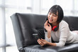 © crizzystudio - Portrait of a happy young Asian woman listening to music with wireless headphones from a music application on a mobile smartphone in the living room at home