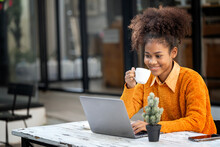 Attractive African Young Confident Woman Holding Coffee Cup And Using Laptop Computer, Sitting At Open Co-workspace.