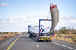 Powerful big rig semi truck with additional axle dolly with oversize load sign transporting wind turbine blade running on the straight highway road