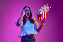 Portrait Of Young Emotive Girl Posing In 3D Glasses With Popcorn Basket Over Pink Background In Neon Light. Concept Of Emotions, Youth, Lifestyle