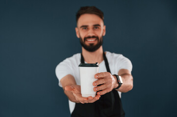 Wall Mural - Giving the cup of coffee. Cafe worker is in apron. Handsome man is in the studio against blue background