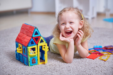 Portrait of a laughing girl on the carpet of the house next to a toy house made of magnets.