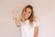 © Марина Демешко - A young attractive caucasian smiling cheerful blonde woman in a white t-shirt shows a peace gesture with her hand isolated on a beige background. Girl shows a victory sign