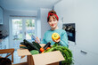 © okrasiuk - Online home food delivery. Woman checking her online order list on her phone. Cardboard box with fresh vegetables and fruits standing on the kitchen table. Local farmer food. Start of a healthy life.