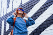 © okrasiuk - Stylish woman in blue jacket and heart shaped sunglasses wearing wireless headphones and listening to music while walking near city urban painted wall. Fashionable hipster lifestyle. Selective focus