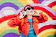 © okrasiuk - Emotional stylish woman in bright clothes wearing wireless headphones, listening to music and singing on rainbow graffity on the brick wall background. Fashionable hipster lifestyle