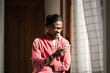 © DimaBerlin - Smiling young black man holding cellphone, standing near window. Happy content African American guy looking at mobile phone screen surfing internet chatting having fun scroll web pages read funny news
