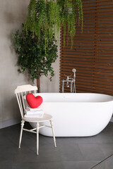 Modern bathroom interior design with concrete walls, wooden slats and white bathtub. vintage chair with towels and heart. Valentine's day concept, relaxation, massage, gifts, care, rest.